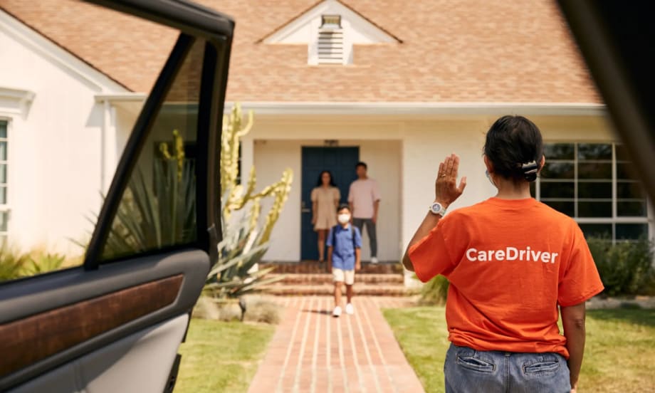 A HopSkipDrive caregiver waits by an open car door in front of a house.
