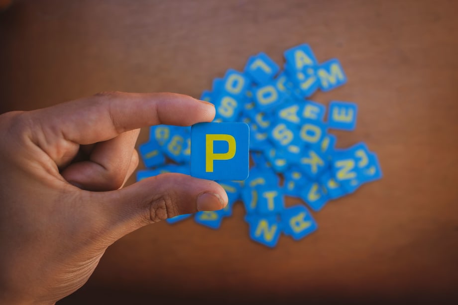 Photo of man's hand holding a square blue piece with a yellow capital P on it, with other letter pieces below on a surface