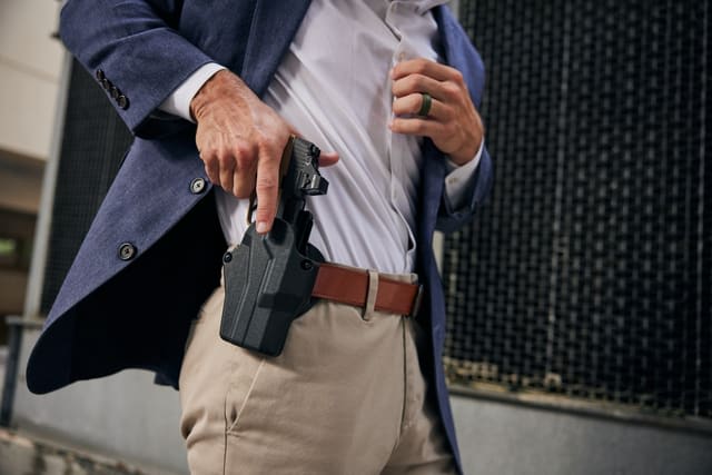 Man in a blue blazer lifting his shirt to reveal a concealed handgun in a waistband holster while standing outdoors.