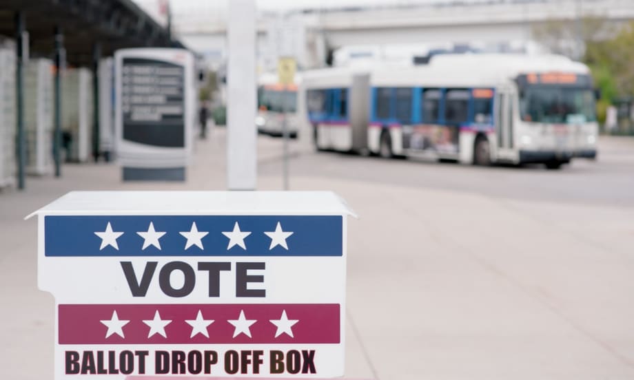 A ballot drop-off box marked “Vote” stands in the foreground at a transit station, with public transit buses visible in the background.