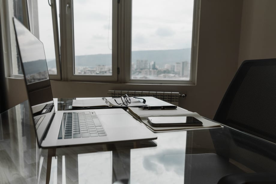 Photo of a laptop, cellphone and eyeglasses on a desk next to an office window