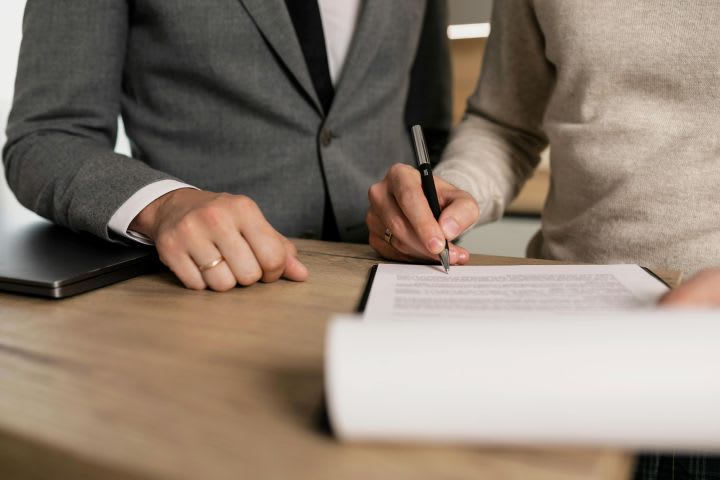 Photo of a man signing a paper at a desk while a man in a suit looks on
