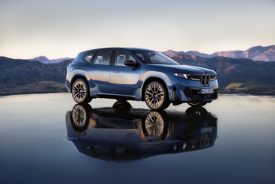 Blue BMW iX3 electric SUV parked on a reflective surface with mountain scenery in the background at dusk.