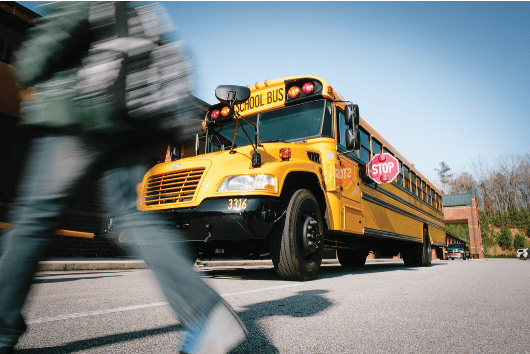 A yellow school bus with its red stop arm extended is stopped to load or unload students as a blurred child crosses in the foreground, illustrating the importance of motorists stopping for school buses and protecting students in the loading zone.