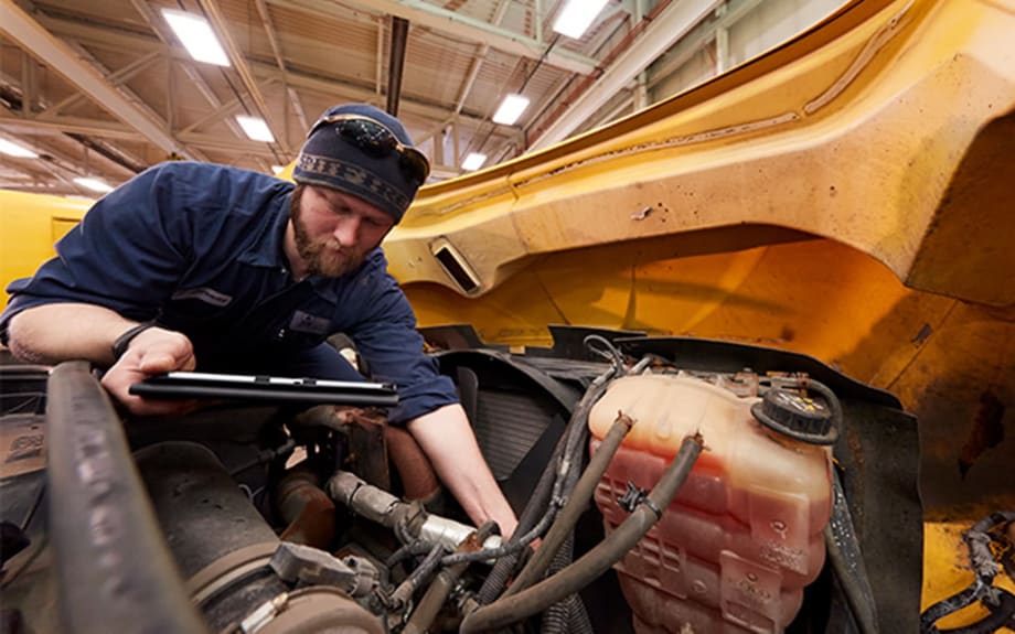 A mechanic in a workshop leans over the open engine compartment of a large yellow vehicle, inspecting components while holding a tablet.
