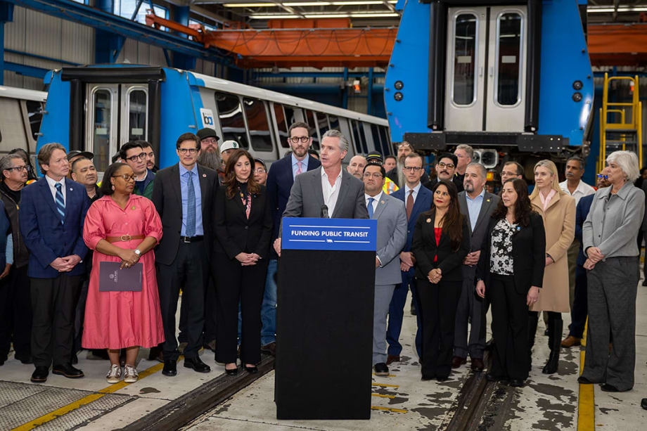 California Governor Gavin Newsom stands at a podium surrounded by a group of state and community leaders.