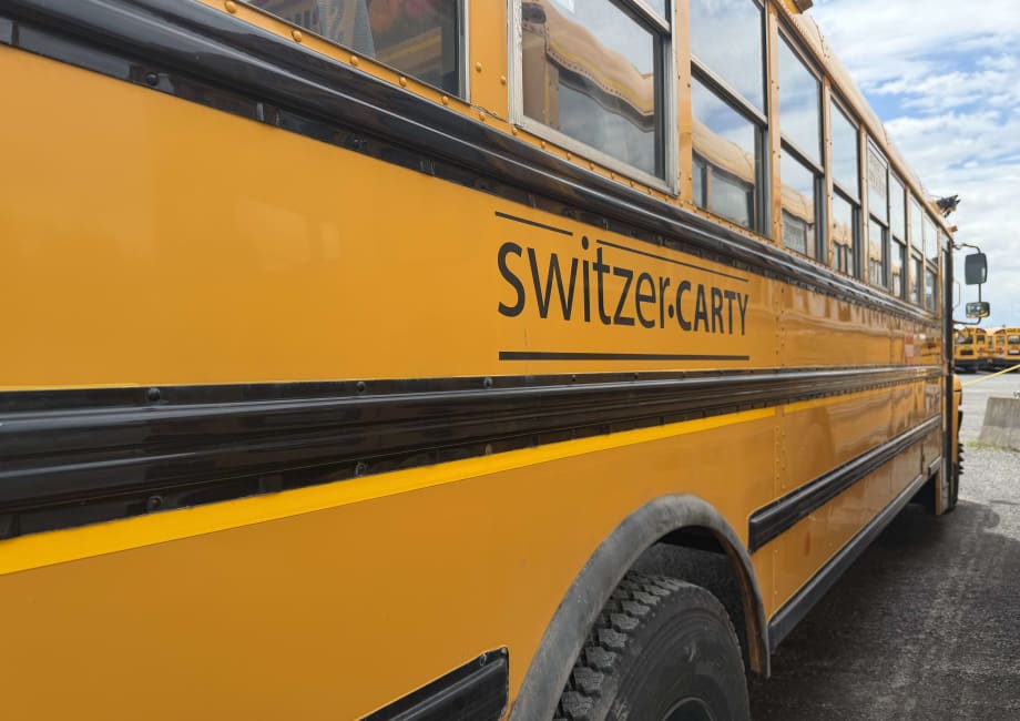 A close-up side view of a yellow Switzer-CARTY school bus parked in a bus yard, with the contractor's name displayed on the side panel and additional school buses visible in the background.