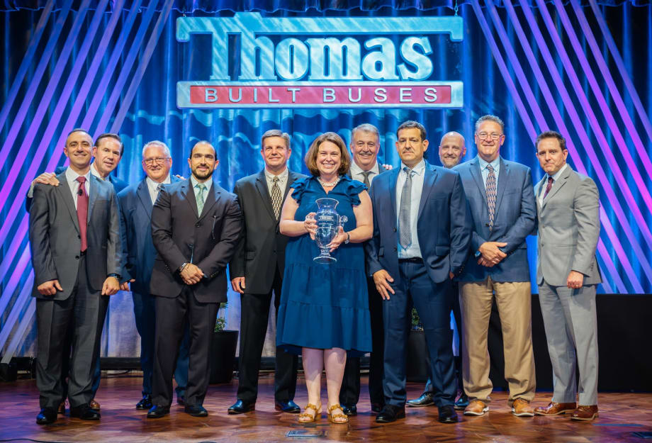 Group of Thomas Built Buses executives, employees, and award recipients pose onstage during an awards ceremony beneath a large Thomas Built Buses sign, with one woman holding a glass trophy at center stage.