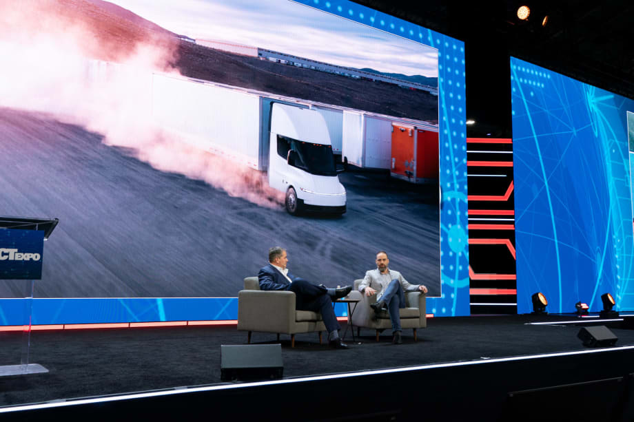 Two men in chairs on stage with big video screen behind them showing Tesla Semi