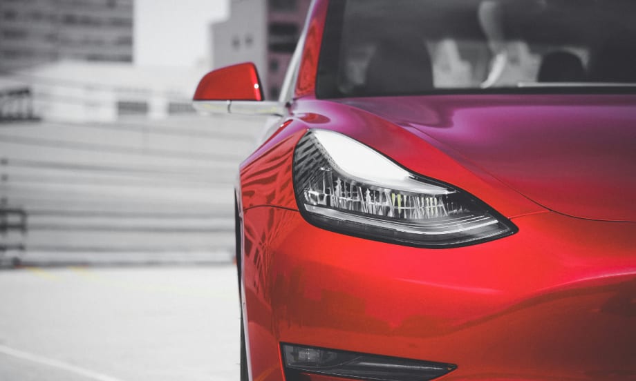 A close up of the right front of a red Tesla in front of a blurred cityscape background.