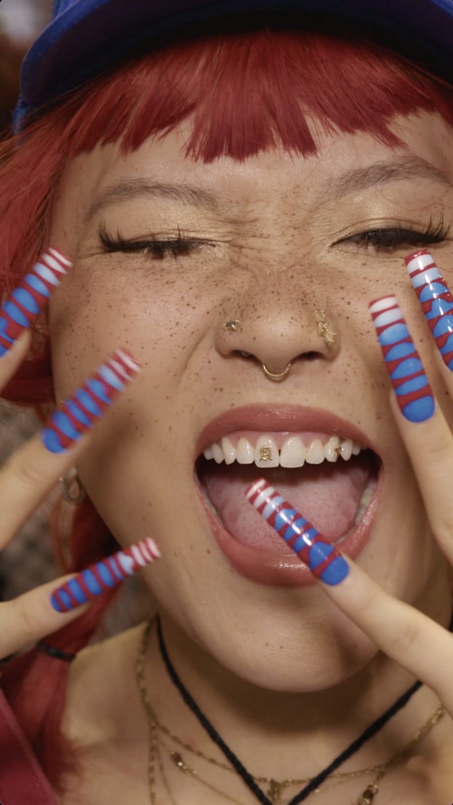 The close up of a young woman with red hair and long, colorful nails that she is holding close to her face.