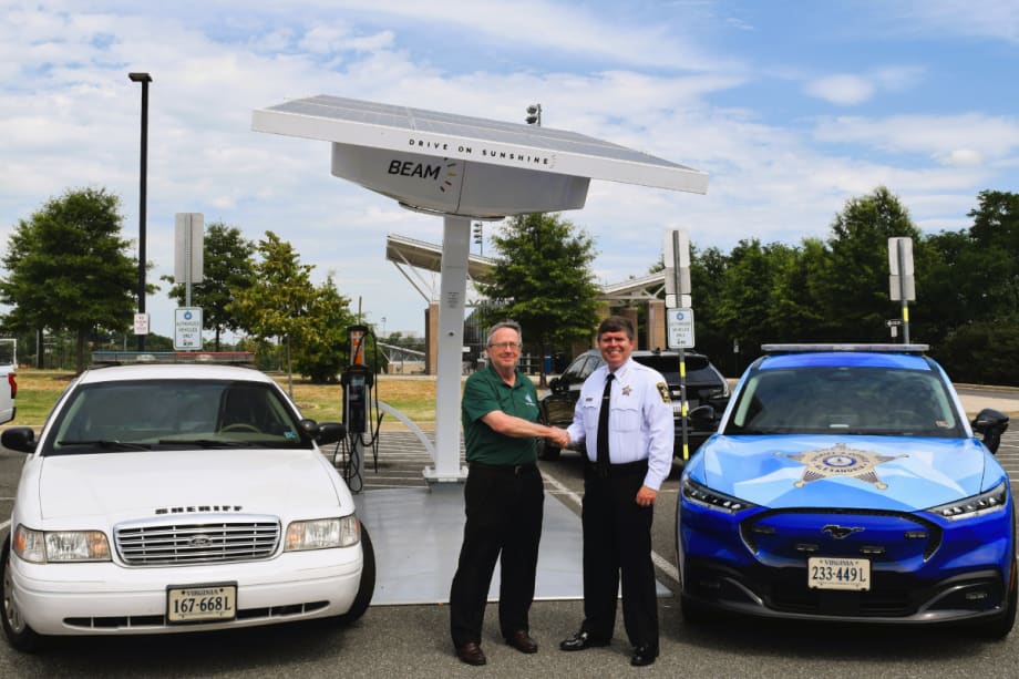 A white Ford Crown Victoria Police Interceptor and blue Mustang Mach-E are shown sitting in front of a Beam solar charger. The fleet manager and sheriff are seen shaking hands between the vehicles.