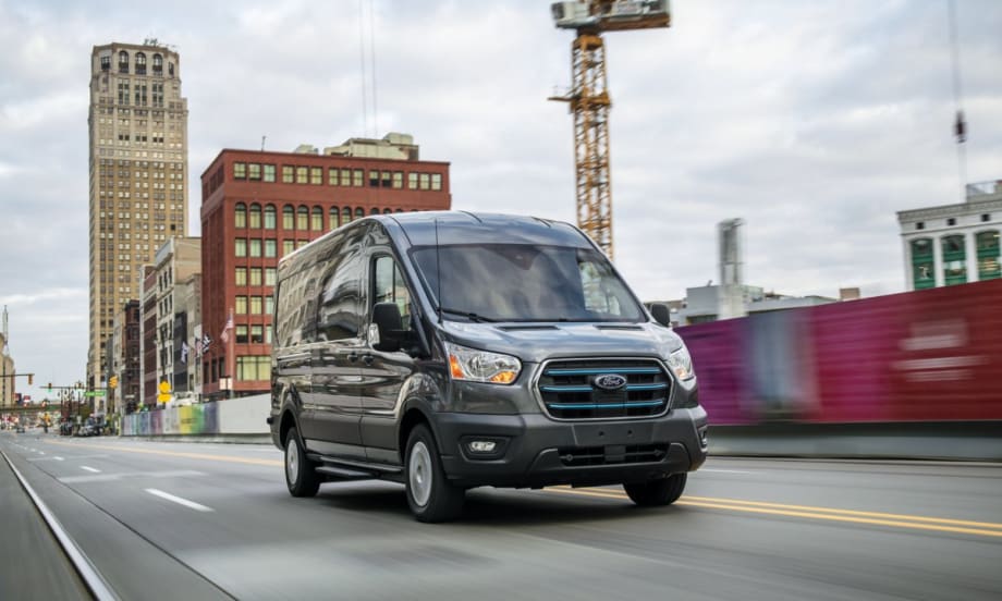 A gray 2022 Ford E-Transit driving on a city road with construction in the background.