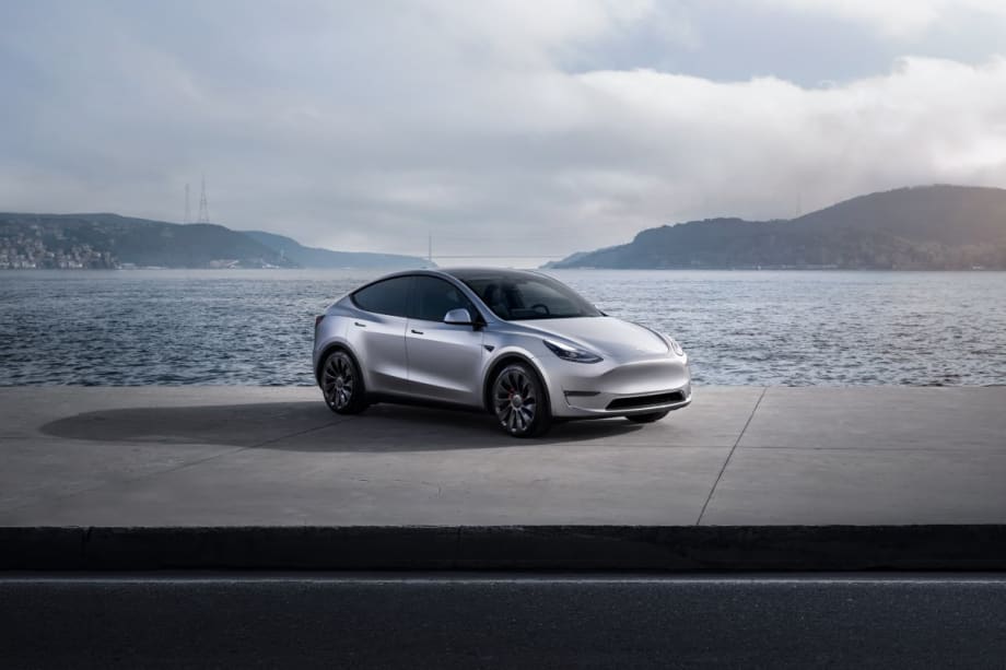 A silver Tesla Model Y parked in front of a body of water and hills in the background.