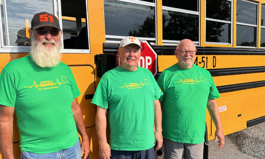 Three men in green shirts pose next to a school bus.