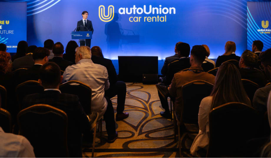 AutoUnion COO speaks to audience in front of wide blue screen with company logo on stage.