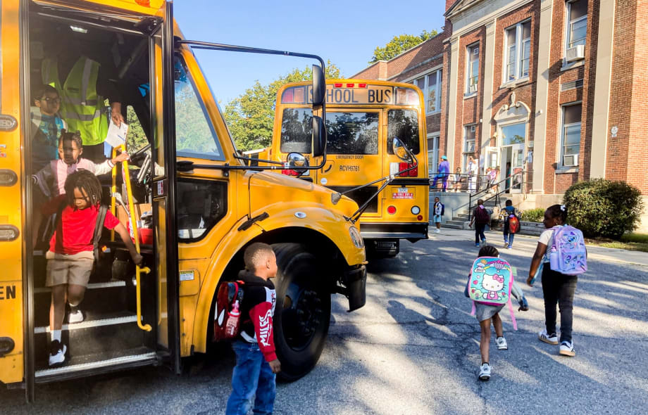 Students with backpacks exit school buses and walk toward a school building as transportation staff assist with afternoon drop-off.