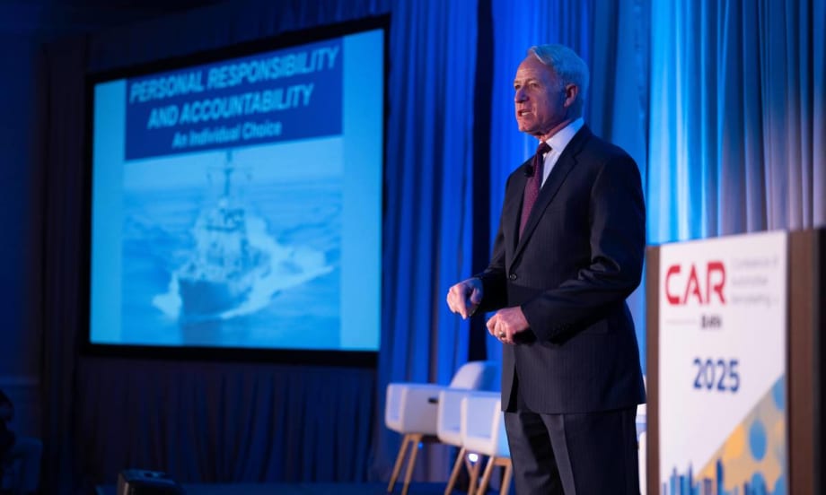 Commander Lippold on stage in front of projected photo of USS Cole.