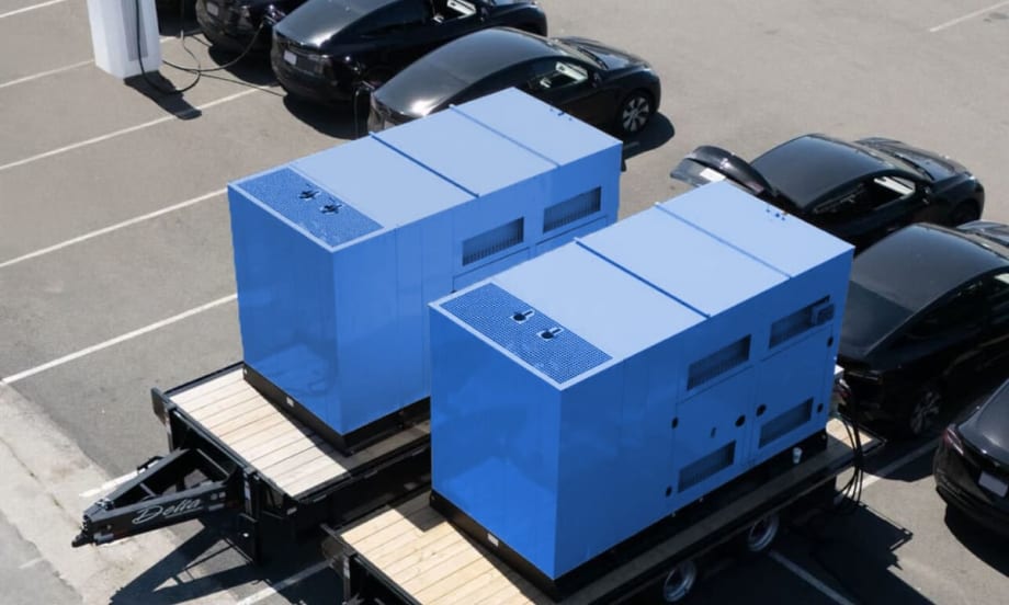 Two blue large box mobile chargers parked along a row of black Tesla EV sedans in a parking lot.