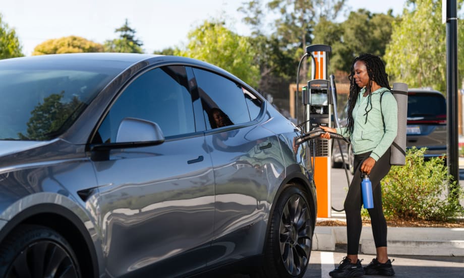 Person charging an electric vehicle at a public EV charging station, holding the charging connector next to a parked electric car outdoors.