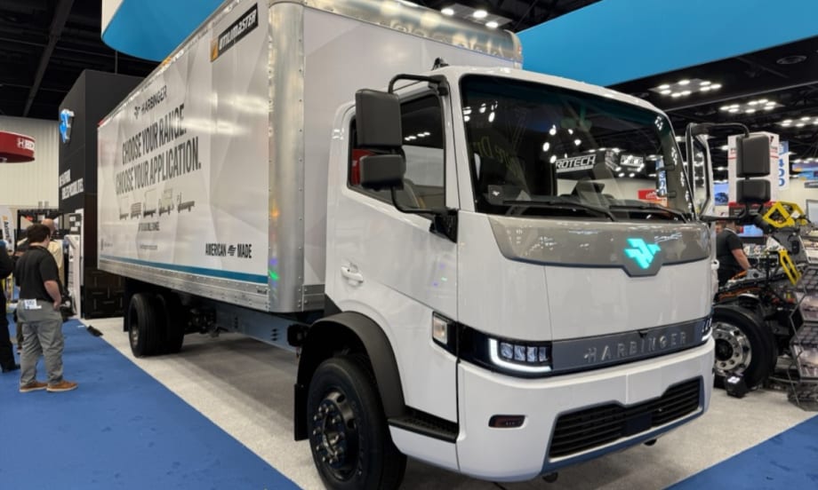 Harbinger HC Series Cab medium-duty electric work truck driving through a city with glass office towers, demonstrating the low cab forward fleet vehicle platform introduced at Work Truck Week.