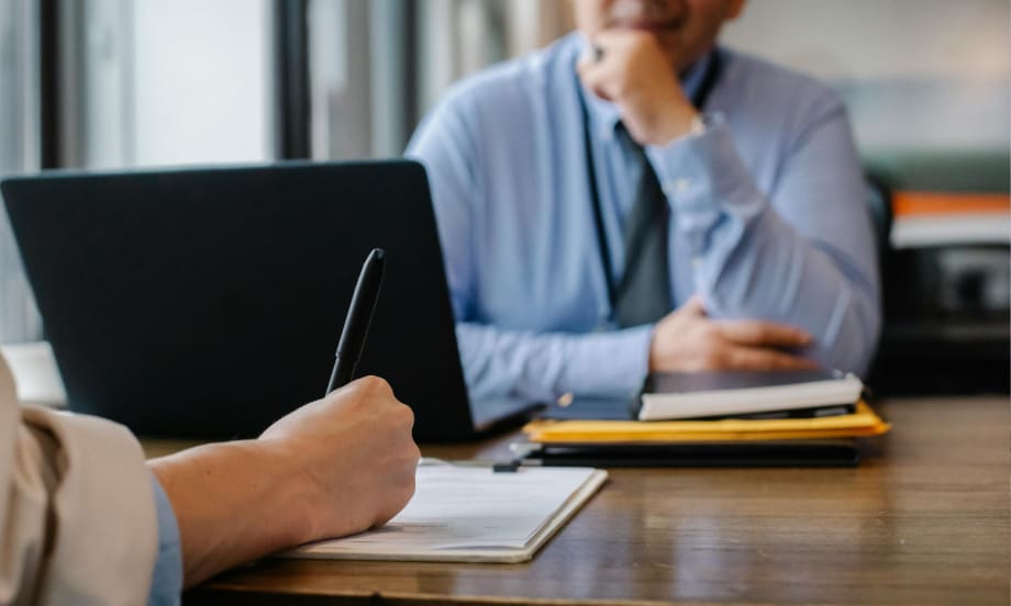 Two people facing each other at a desk, the person in the foreground is filling out papers.