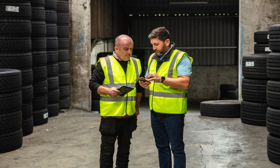 Two workers in safety vests look at a tablet and phone in a tire warehouse.