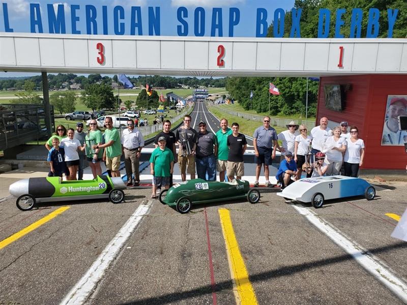 A group of participants and organizers stand behind three colorful soap box cars at the starting line of the All-American Soap Box Derby track in Akron, Ohio, celebrating the launch.