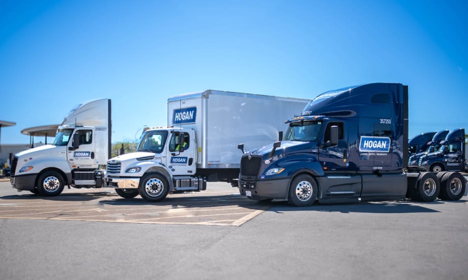 Three Hogan semi-trucks sit in a row in a parking lot on a sunny day.