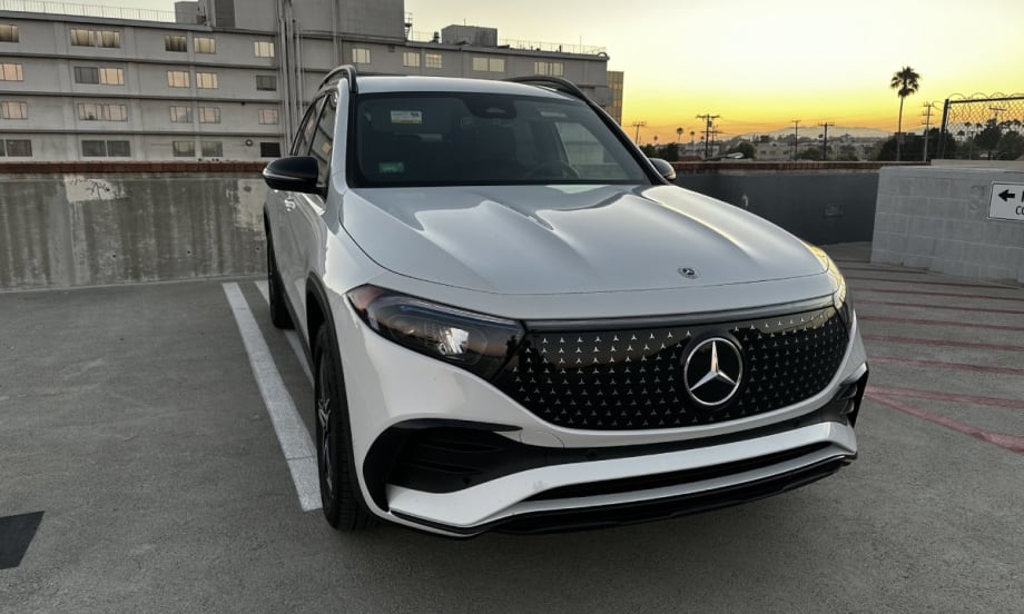 The front of a white Mercedes-Benz EQB 250+ in a parking space with a sunset in the background.