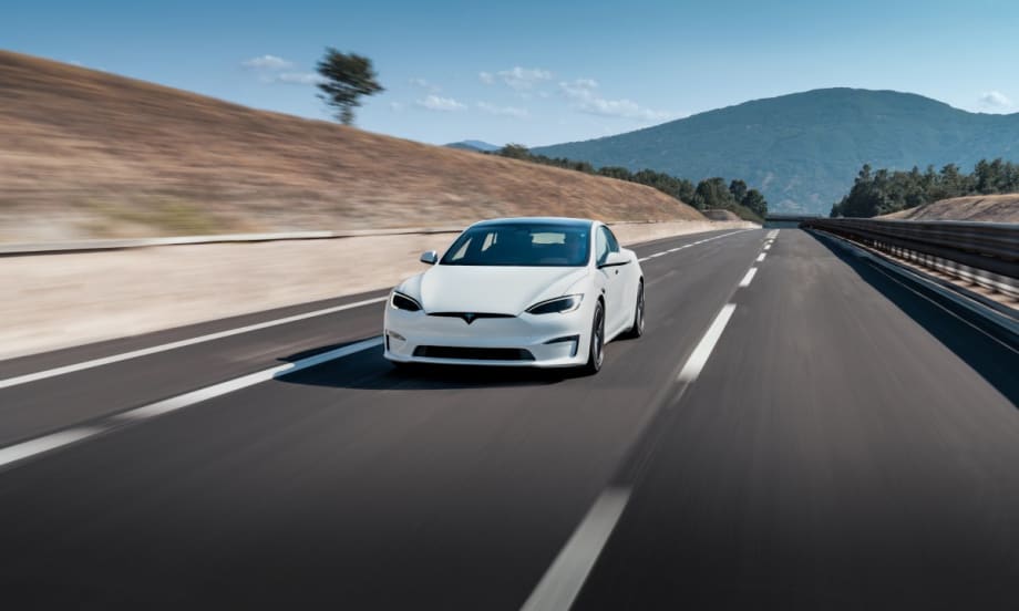 A white Tesla Model S driving down a paved road with hills and mountain in background on a sunny day.