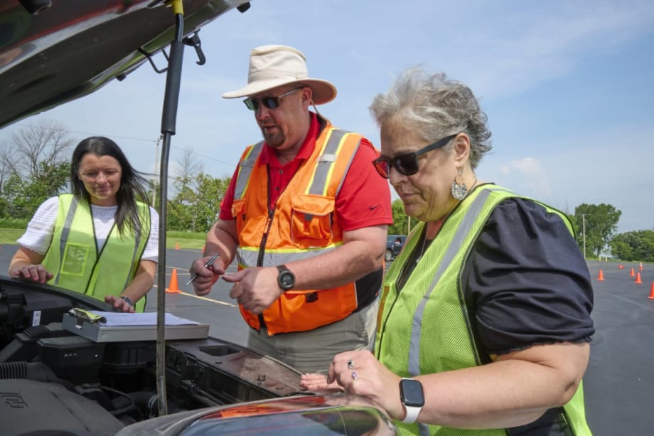 A man in an orange vest shows two women in green vests items under the hood of a vehicle.