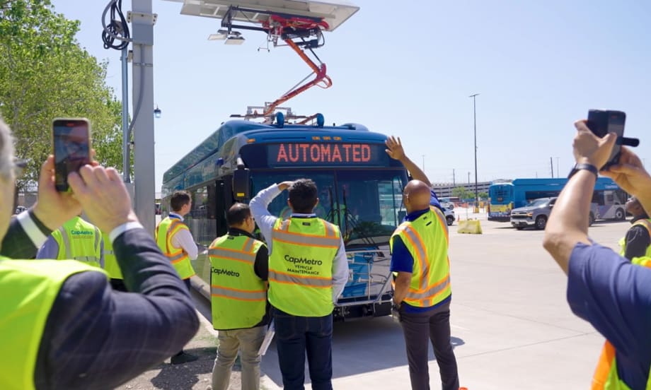Image of a blue CapMetro bus at an overhead charging station as employees watch.