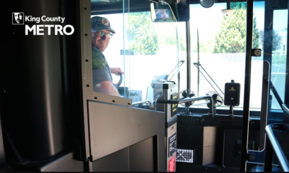 Image of a bus operator posing from the front seat of a public bus, behind a clear partition.