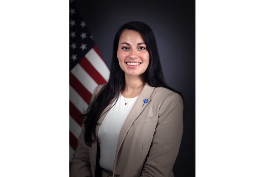 Smiling professional woman in beige blazer standing in front of American flag