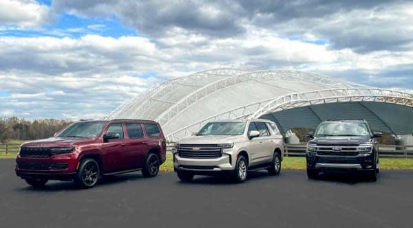 A lineup of large SUVs are shown. From left, those include a maroon Jeep Wagoneer, off-white Chevrolet Tahoe, and navy blue Ford Expedition.