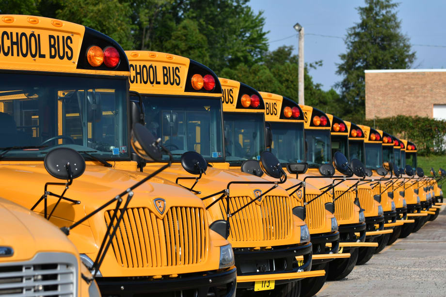 Row of yellow school buses parked in a lot, lined up bumper-to-bumper with mirrors extended, photographed outdoors in daylight with trees and buildings in the background.