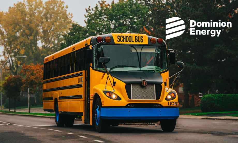 A LionC all-electric school bus drivers down a road with colored leaves in the background.
