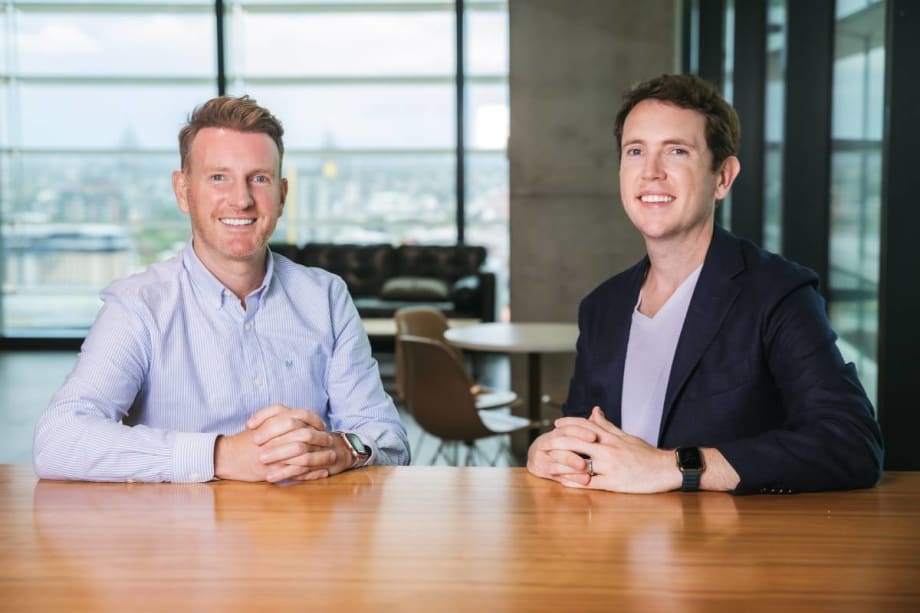 Two corporate-looking young men smile along a gleaming wooden conference table.