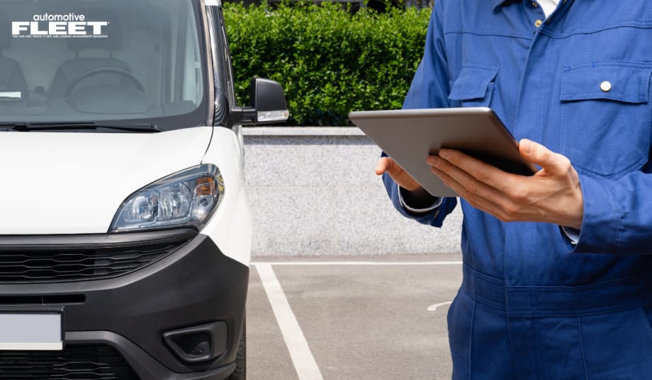 A man in a blue mechanic jumper holds a tablet at chest level in front of a white van.