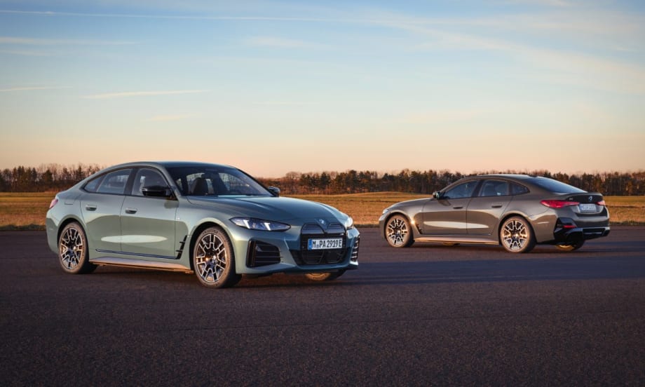 Two 2025 BMW i4 series cars (one light blue and one dark gray) parking on pavement with a field and trees in the background.