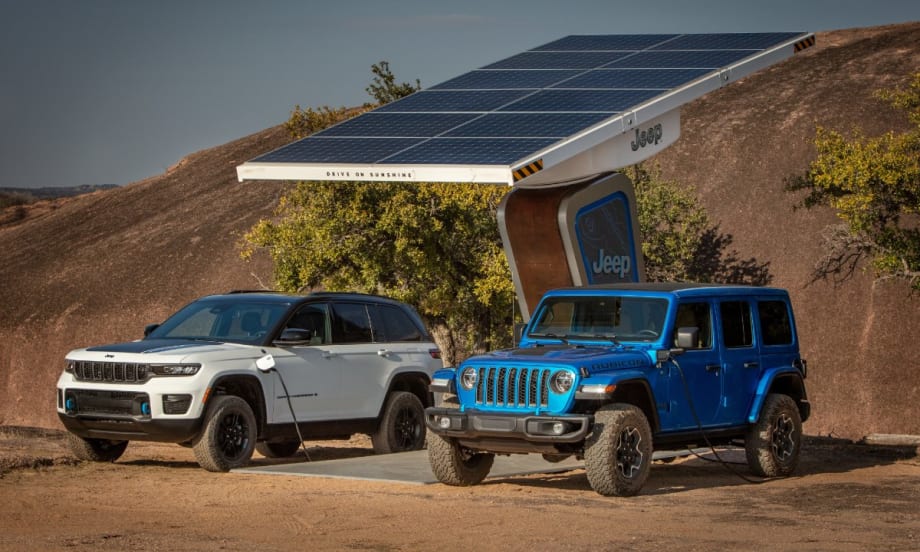A white 2022 Jeep Grand Cherokee and a cobalt blue 2022 Jeep Wrangler recharge at a Jeep charge station in a desert.
