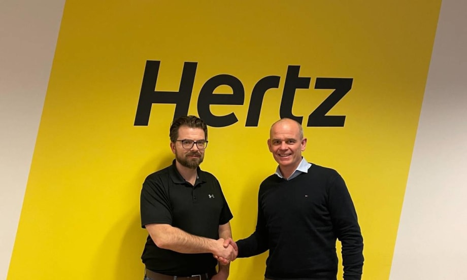 Two executives shaking hands in front of bold yellow Hertz sign.