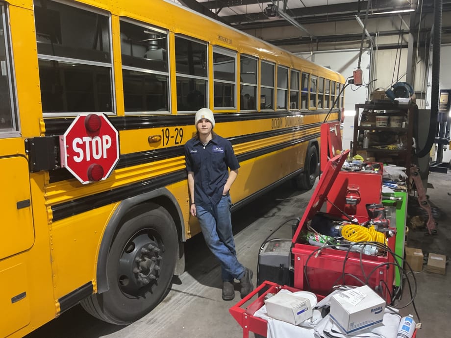 a young man leans against a school bus in a maintenance garage