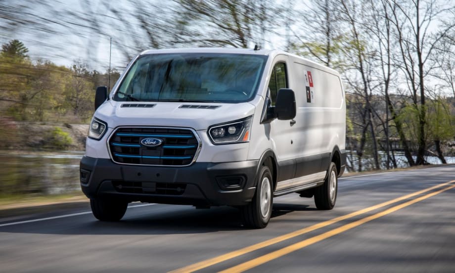 A Ford E-Transit van driving down a road with trees in the background.