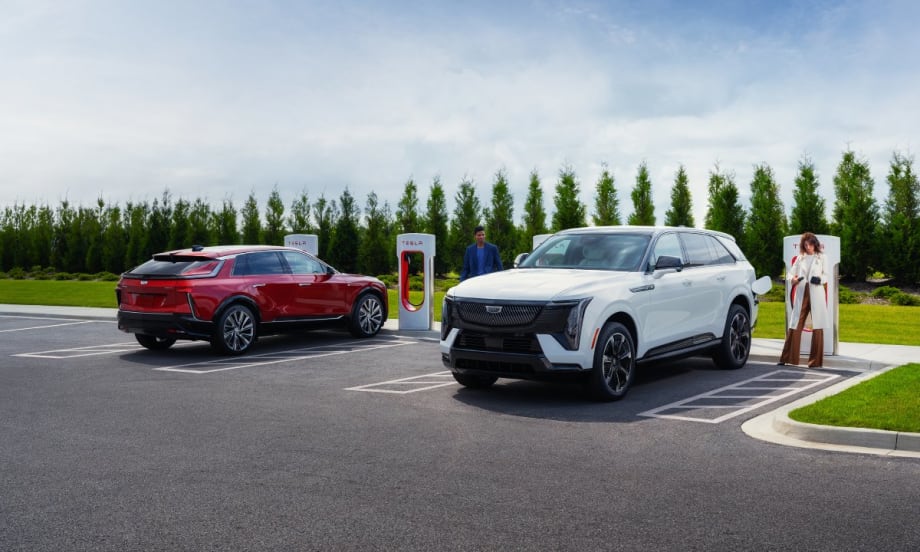 A red suv and a white suv parked in front of tesla superchargers with trees in the background.