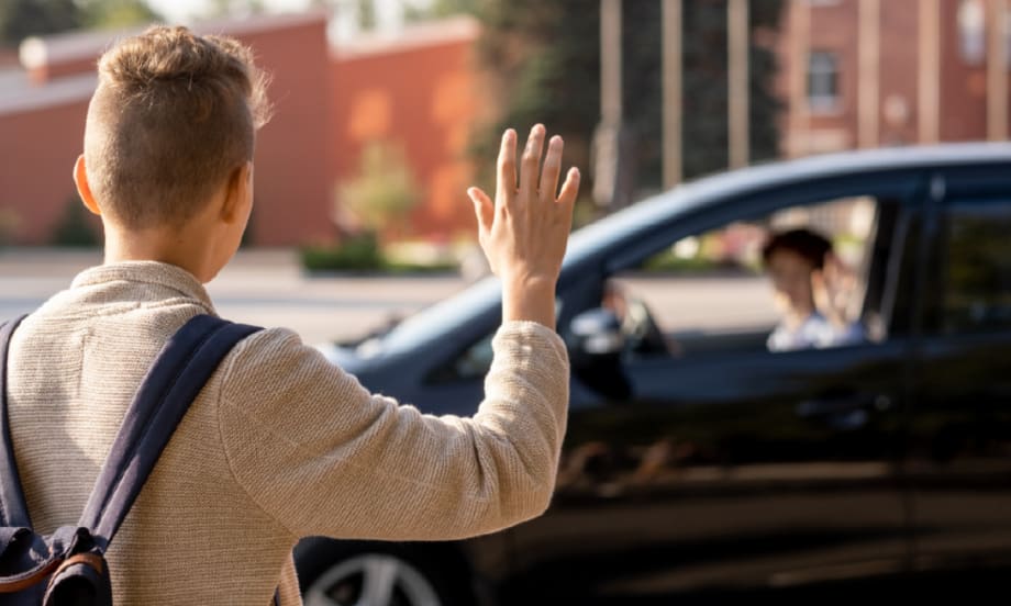A young boy wearing a backpack waves at an adult sitting in a car.