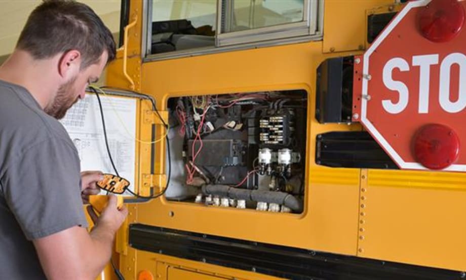 A man in a grey t-shirt works on the electrical components to a school bus stop arm.