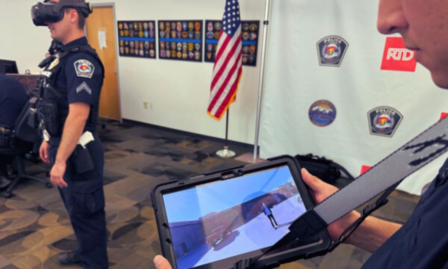 A RTD transit police offer holds up a tablet showing virtual reality training while an officer in the background stands with a VR headset on.