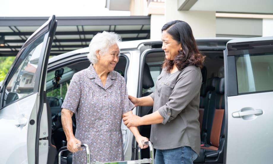 A woman helps an elderly woman with a walker out of van.